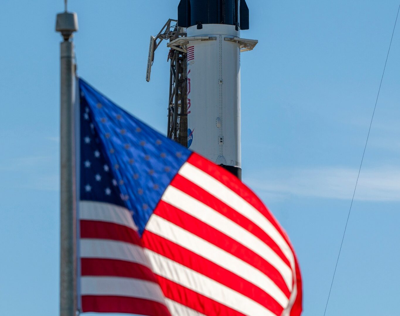 A close-up view of a SpaceX Crew Dragon spacecraft mounted atop a Falcon 9 rocket on the launch pad. The capsule and trunk are connected to ground support hardware, with NASA and mission insignia visible on the rocket’s exterior against a bright blue sky. in the foreground in the American flag.