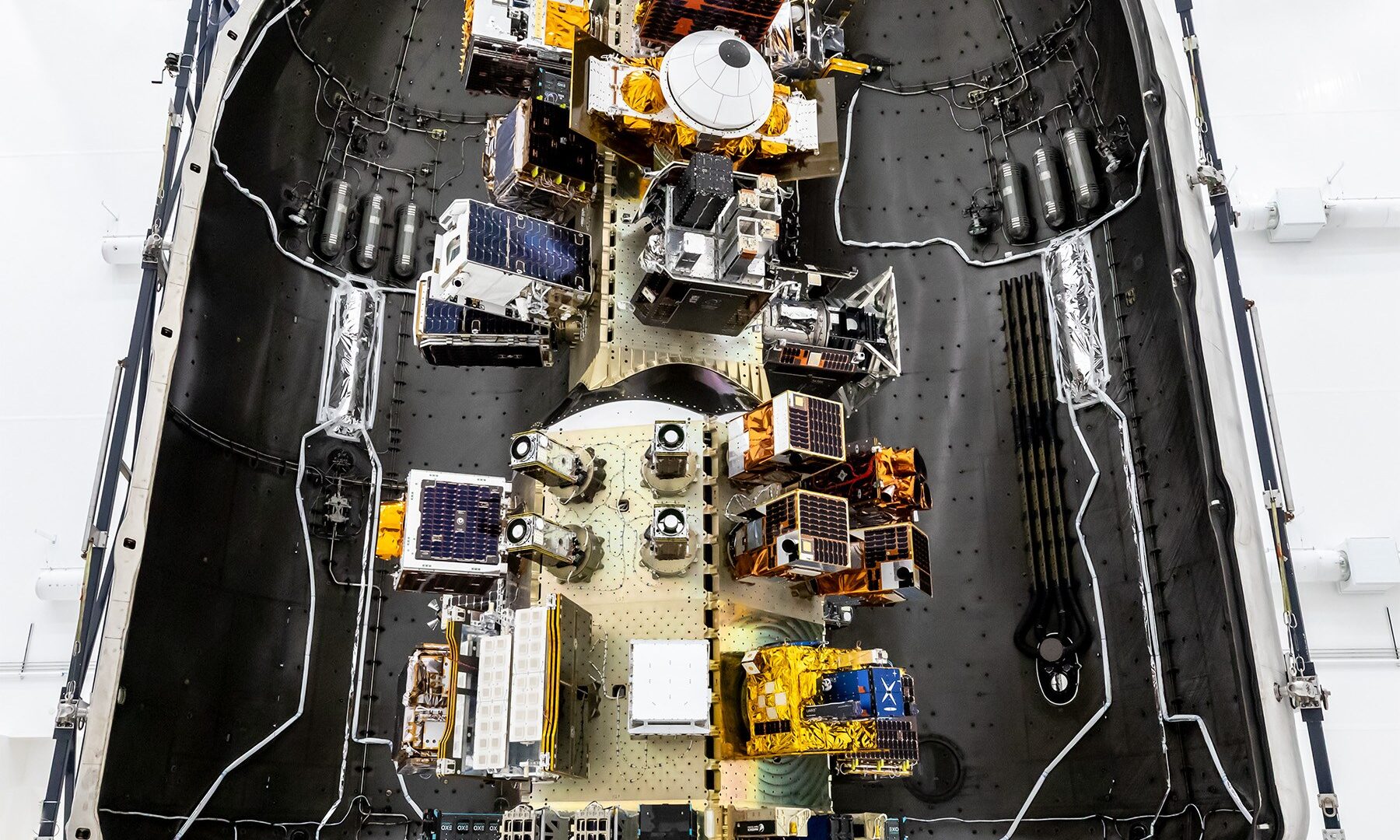 Top‑down view of a SpaceX payload fairing interior showing mounted satellites and hardware arranged along the center structure, surrounded by the curved black fairing walls.